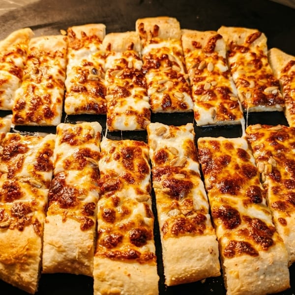 Rectangular slices of cheesy flatbread with sunflower seeds, arranged on a dark serving tray.