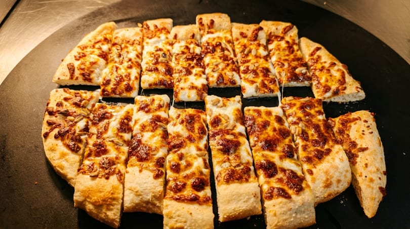 Rectangular slices of cheesy flatbread with sunflower seeds, arranged on a dark serving tray.