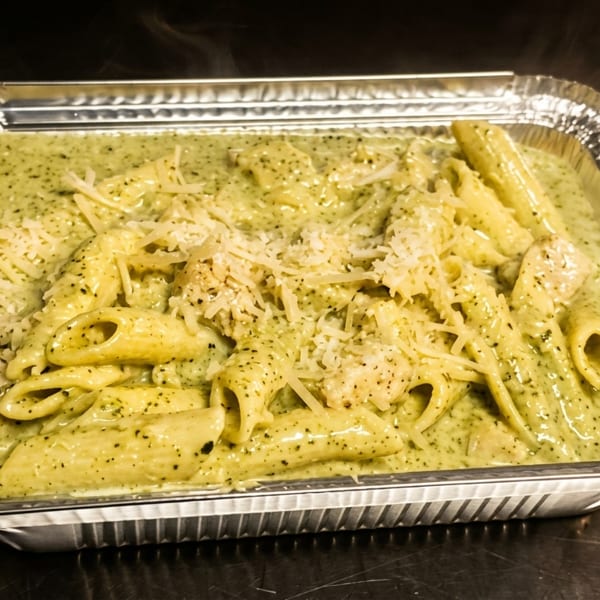 Steaming penne pasta with green pesto sauce and grated cheese in a foil takeout container.