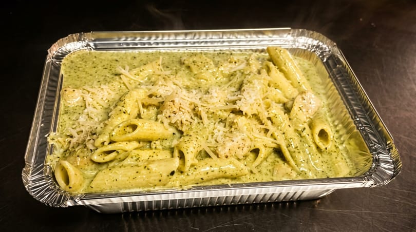 Steaming penne pasta with green pesto sauce and grated cheese in a foil takeout container.